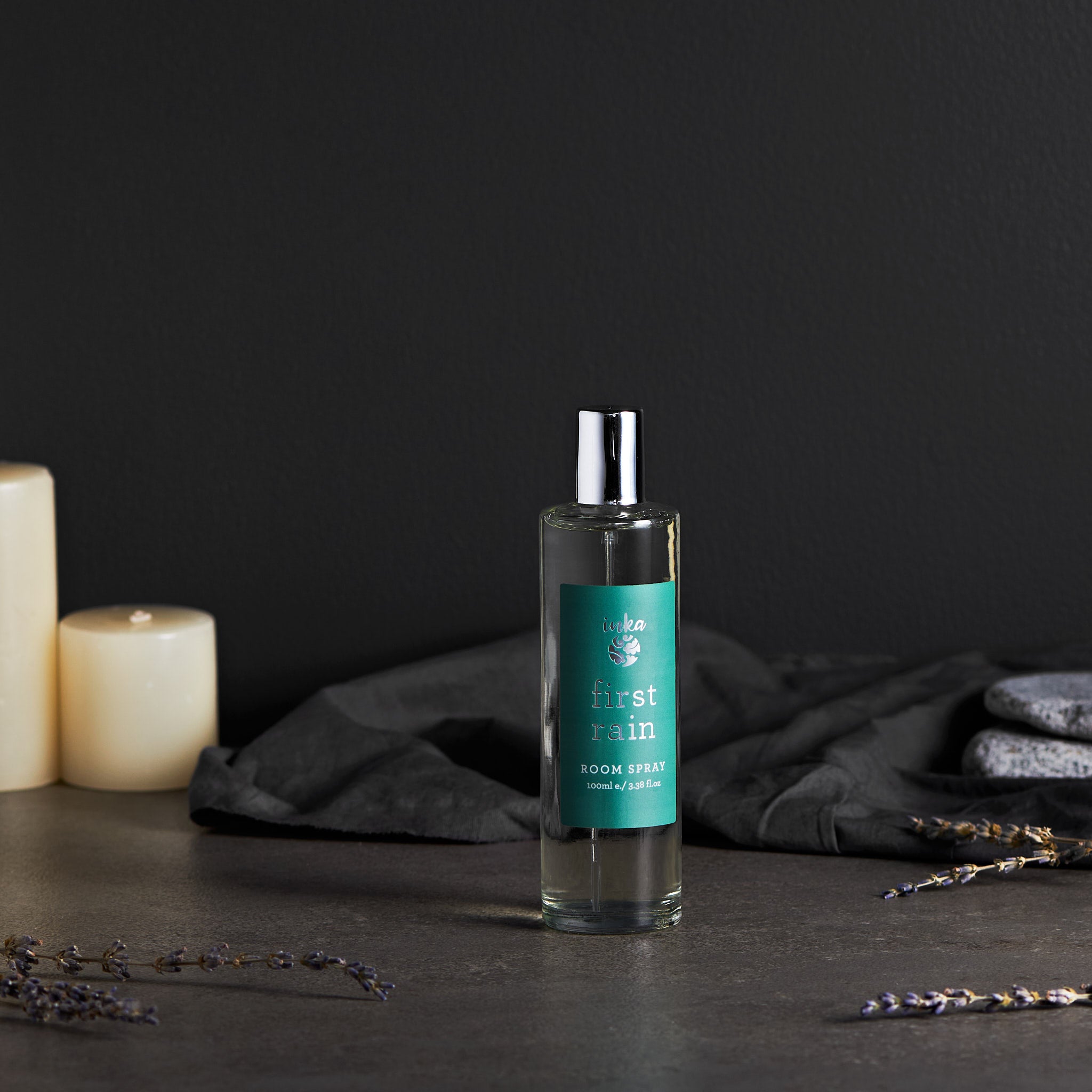 A room spray bottle with a label that reads 'First Rain'. The bottle is clear with a silver cap, placed on a dark surface, with candles and lavender in the background.